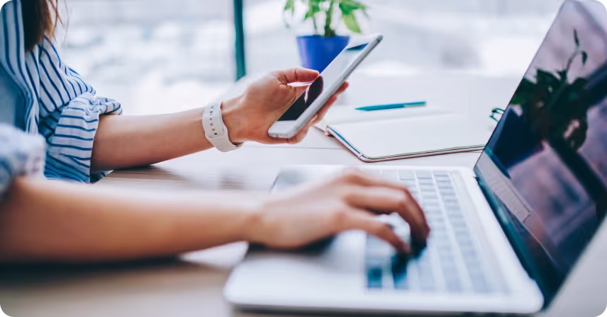 an individual works at a desk, using a smartphone in one hand and typing on a laptop with the other
