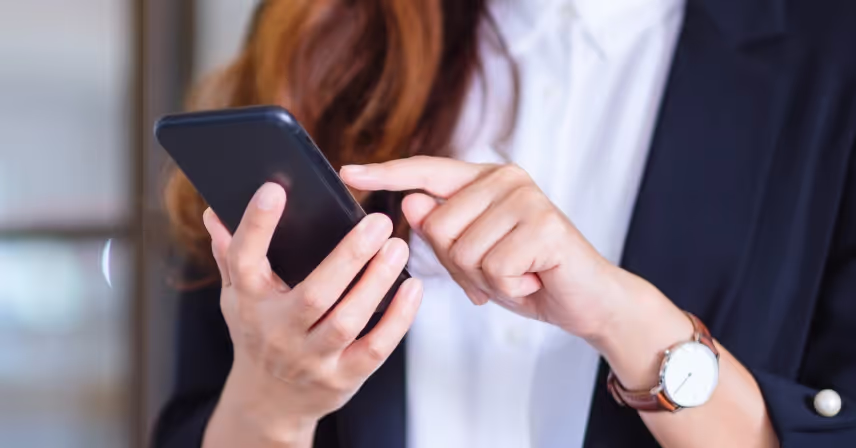 Close-up of a person with long hair wearing a wristwatch, a white shirt, and a dark blazer using a smartphone. The person's face is not visible. They are holding the phone in one hand and navigating it with the other.