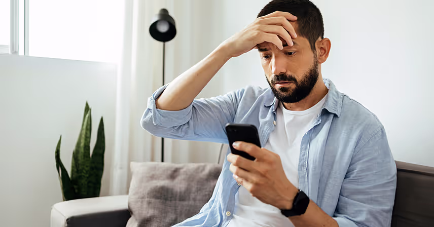 Man wearing sitting on couch looks confused while looking at his smartphone's screen.