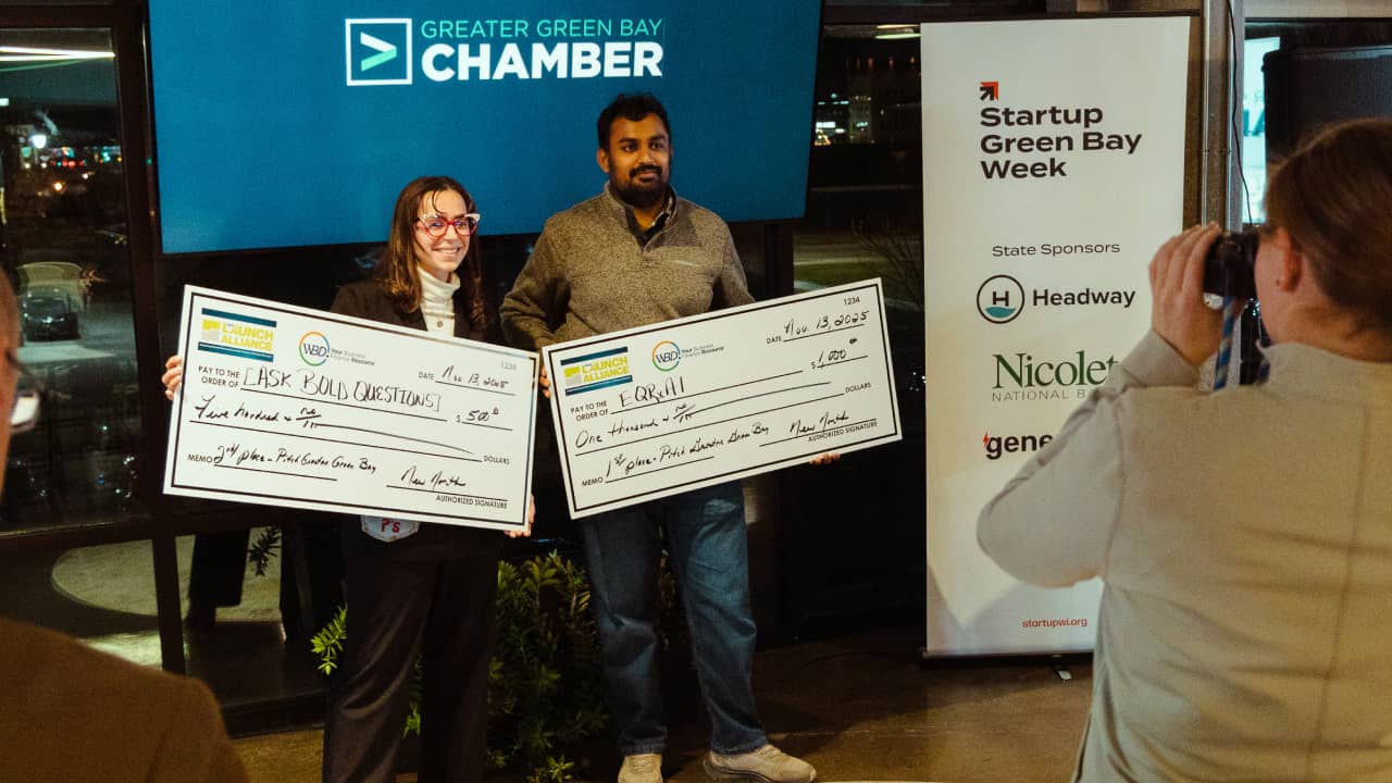 First and second place winners hold oversized prize checks in front of a Greater Green Bay Chamber screen at the Pitch Greater Green Bay competition.