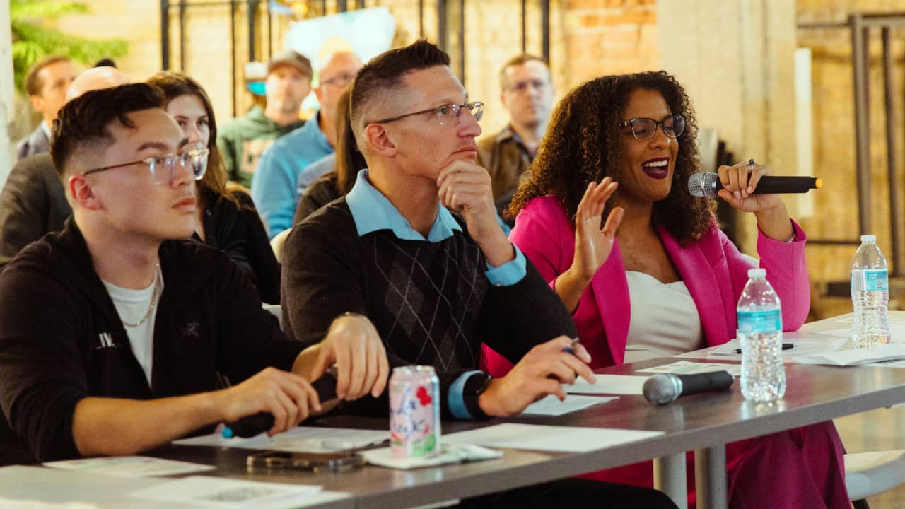 Three judges seated at a table listen to a founder's pitch, one asking a question into a microphone, at a Northeast Wisconsin pitch competition.