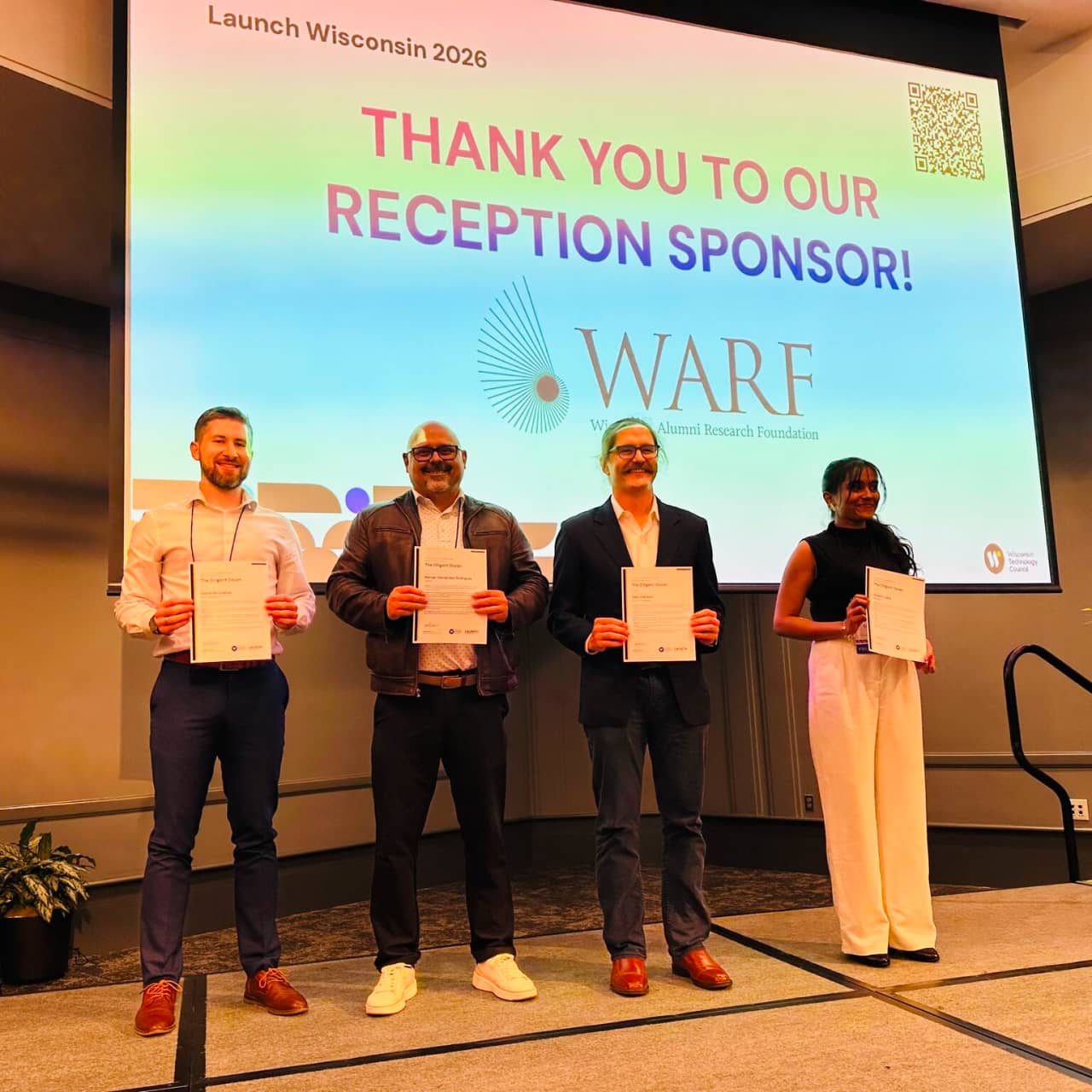 Four Launch Wisconsin 2026 category winners hold certificates on stage in front of a WARF reception sponsor slide.