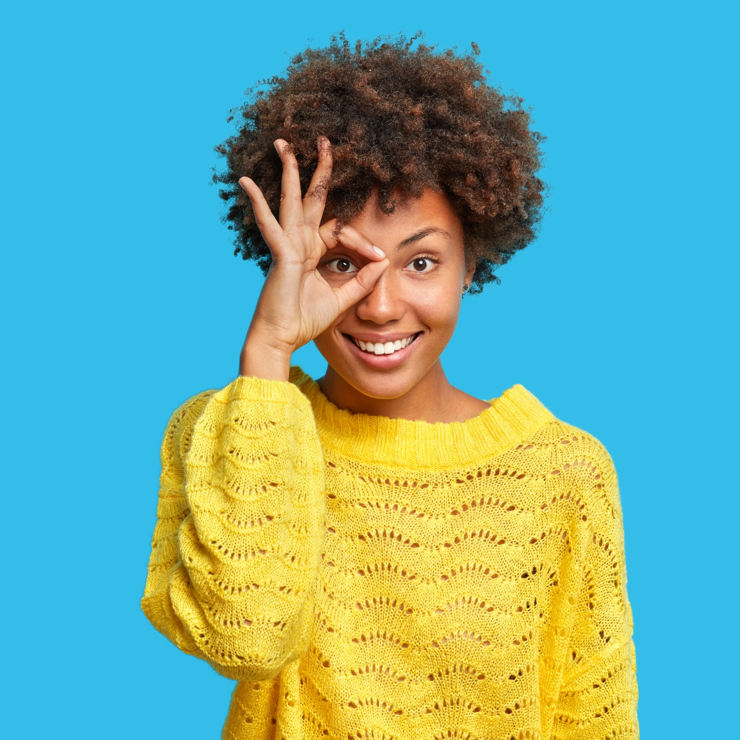 Femme aux cheveux bouclés souriante en pull jaune faisant un rond avec les doigts devant son oeil