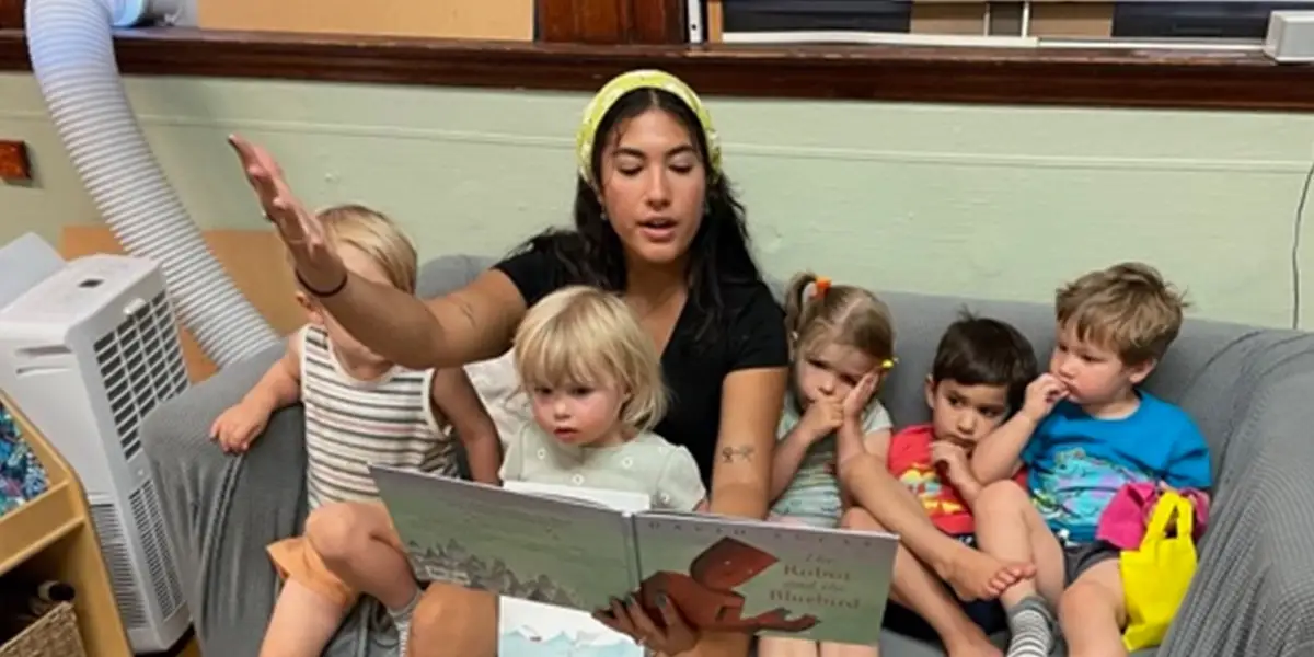A UTCS teacher reads a picture book to a group of toddlers on a classroom couch