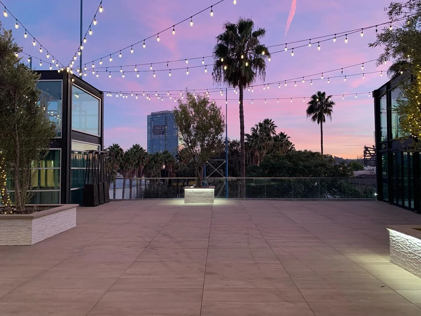 Three-story brick building with large illuminated windows at dusk on a street corner with traffic lights and trees.