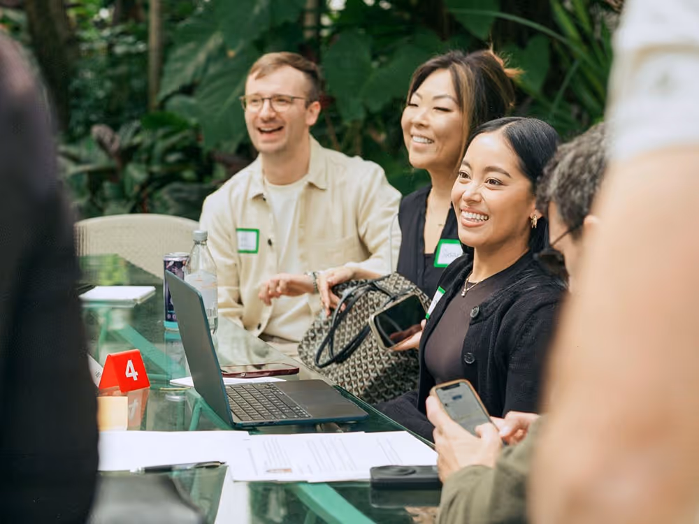 Three people sitting at a glass table outdoors, smiling and engaging in conversation with a laptop and papers on the table.