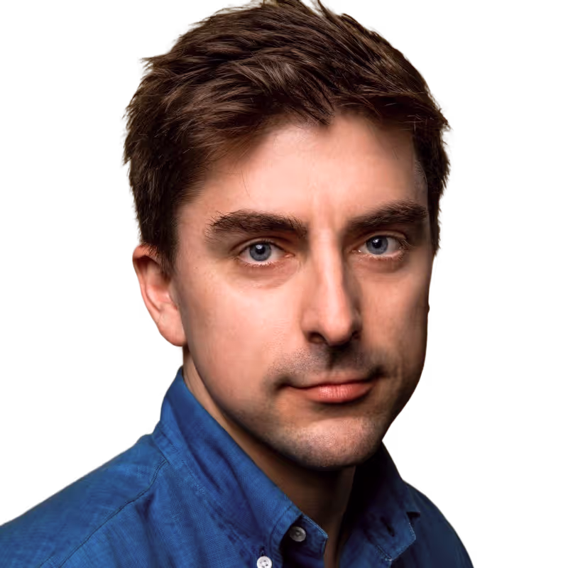 Close-up portrait of Greg Dale with short brown hair and blue eyes wearing a blue collared shirt against a black background.
