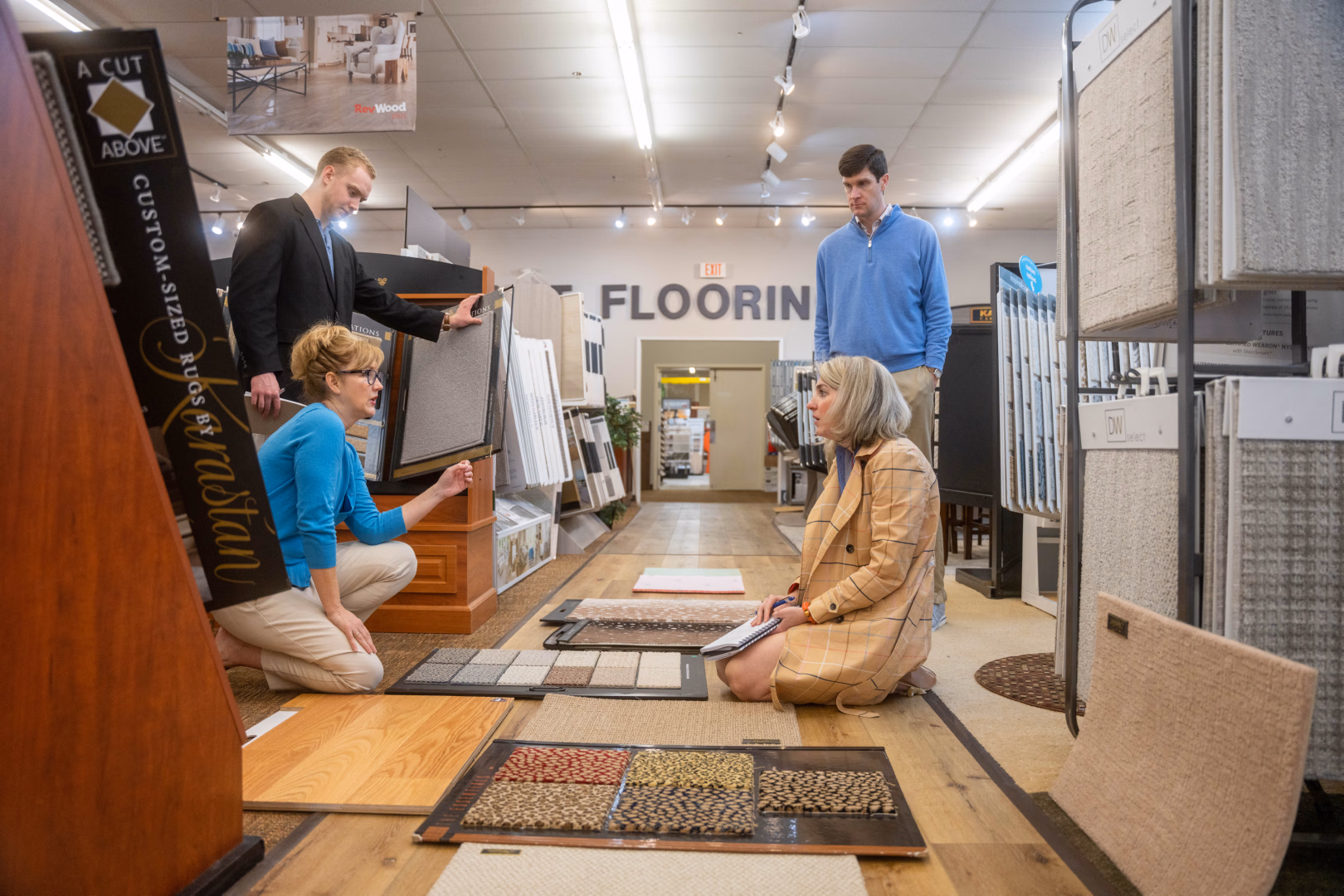 Two women kneeling on the floor examining carpet samples with two men standing behind them in a flooring store.