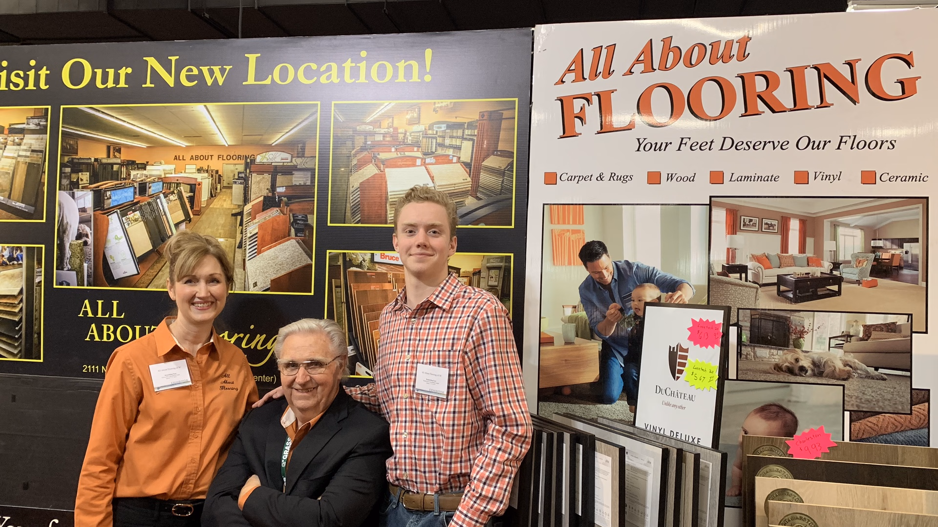 Three people smiling and posing in front of promotional banners for All About Flooring with samples of flooring materials displayed.