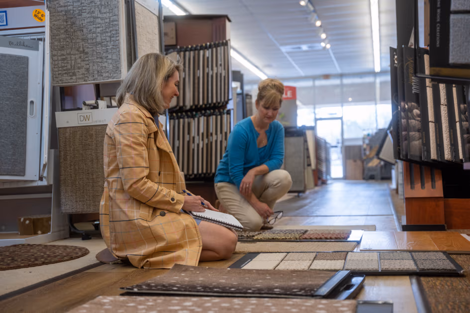 Two women in a showroom examining carpet samples spread on the floor, one taking notes and the other holding glasses.