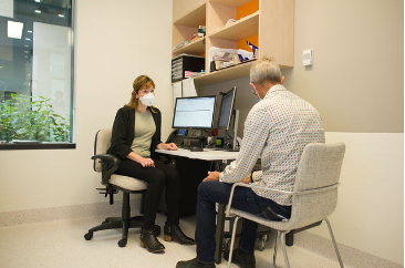 A person wearing a mask sitting at a desk with a person in a chairDescription automatically generated