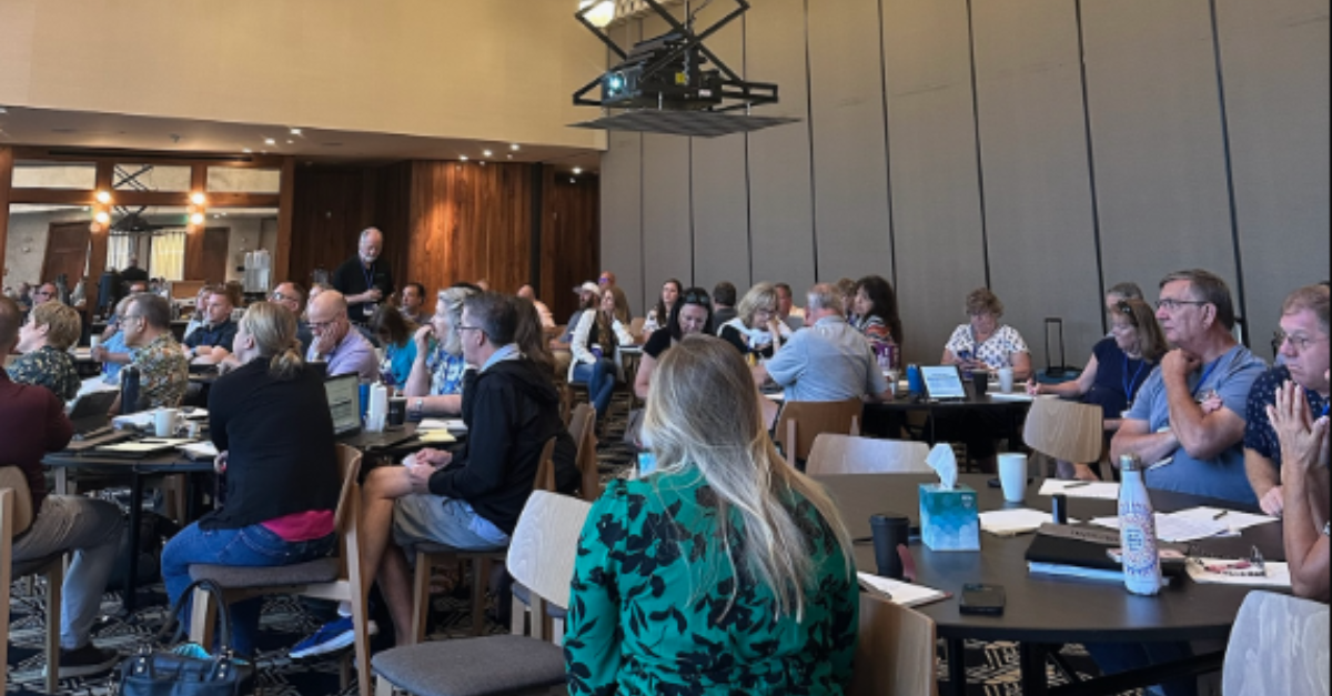 Group of people seated in a conference room, attentively listening to a presentation. Several attendees have laptops and notebooks on the tables.