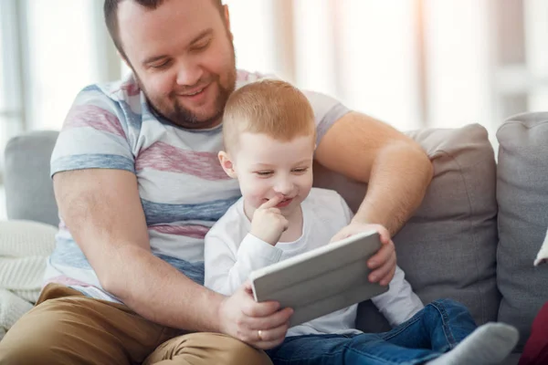 Photo of dad with his son with tablet sitting on gray sofa — Stock Photo, Image