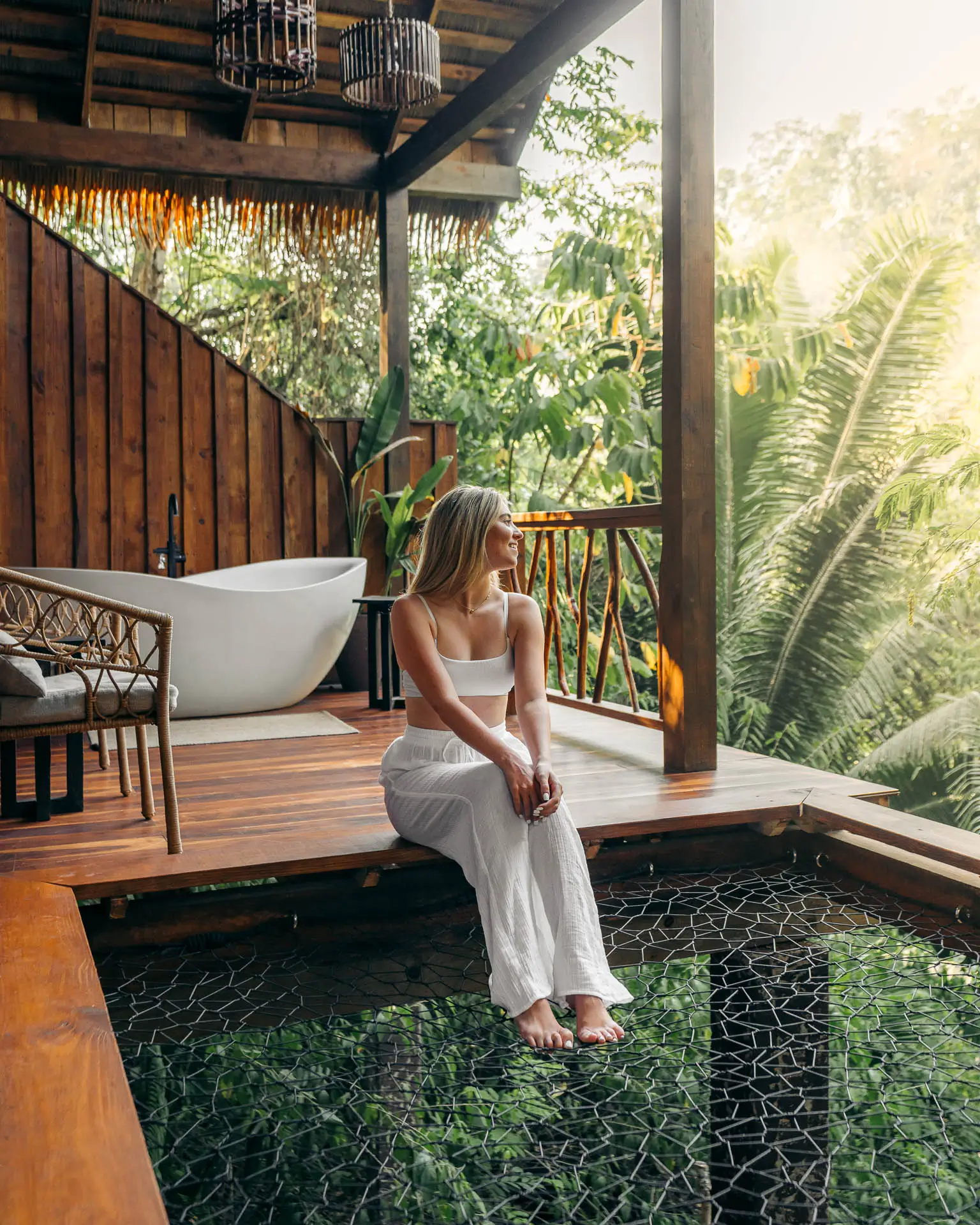 Woman sitting on wood deck with an outdoor net bed, surrounded by tropical plants at Sweet Songs Jungle Lodge in Belize.