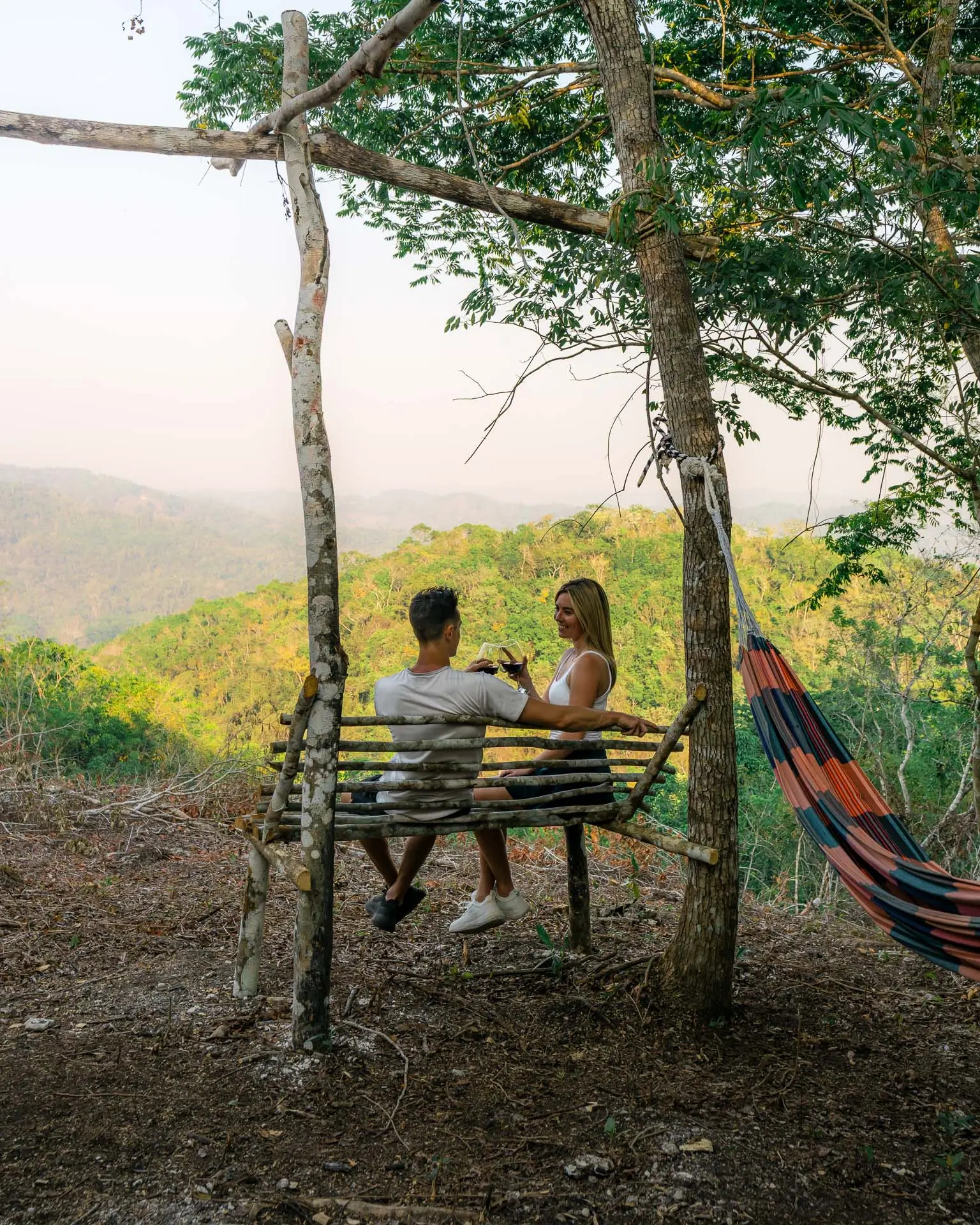 Couple sitting on a rustic wooden bench in a forested area, clinking glasses with a green mountainous view at Sweet Songs Jungle Lodge in Belize.