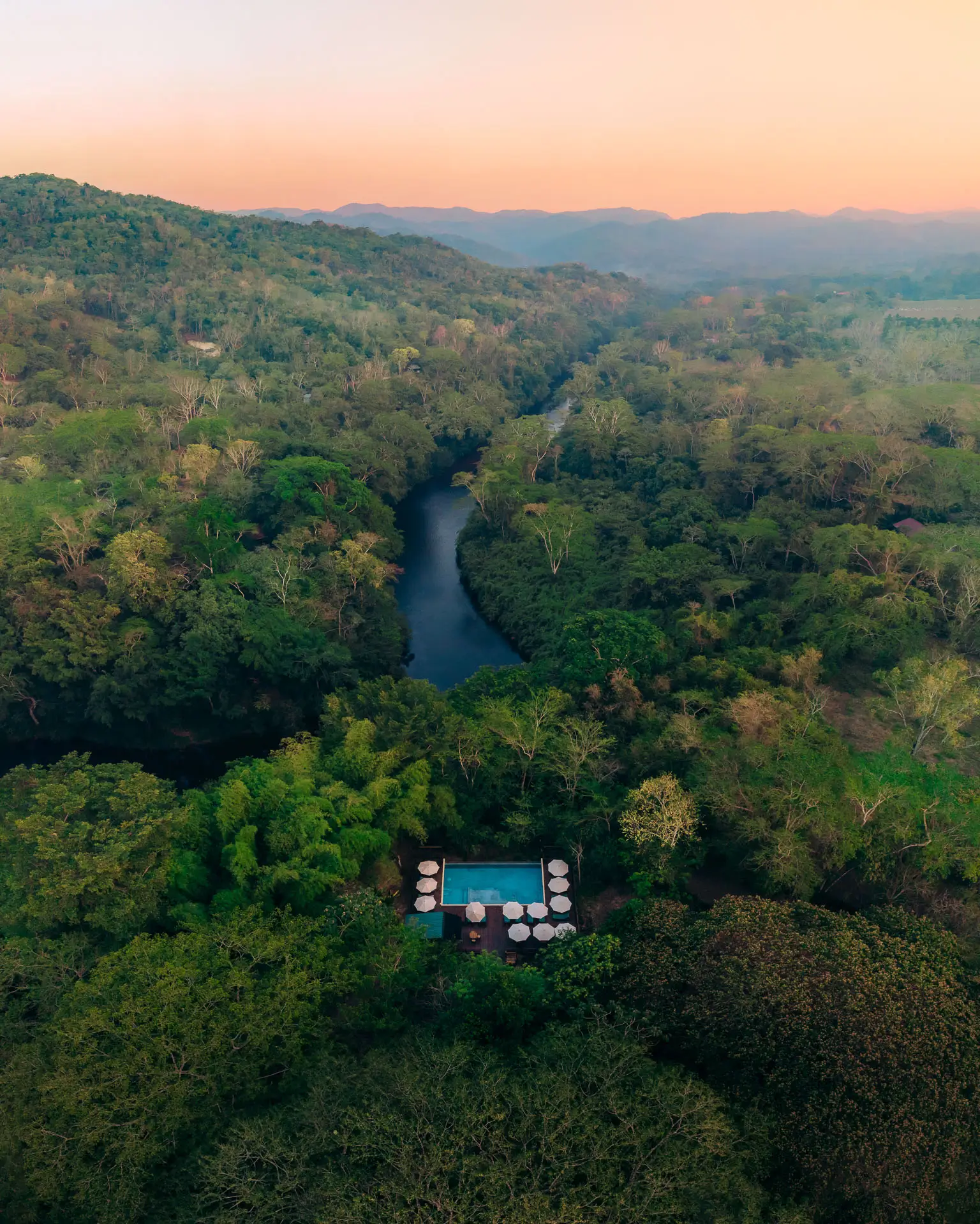 Aerial view of the Macal River winding through dense forest with a swimming pool and umbrellas in the foreground at Sweet Songs Jungle Lodge in Belize.