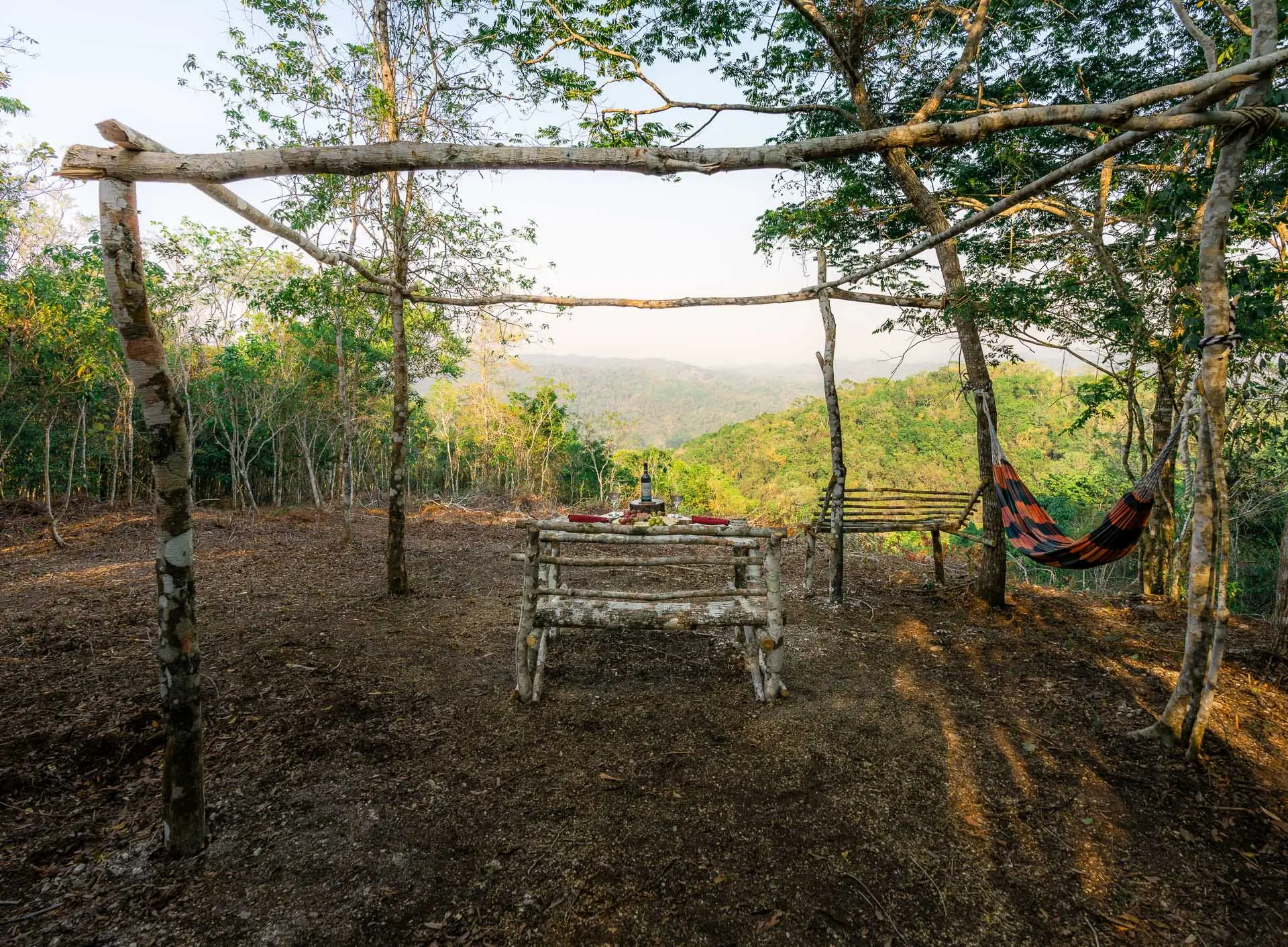 Rustic wooden table and benches under tree canopy with a hammock and forested hills in the background at Sweet Songs Jungle Lodge in Belize.