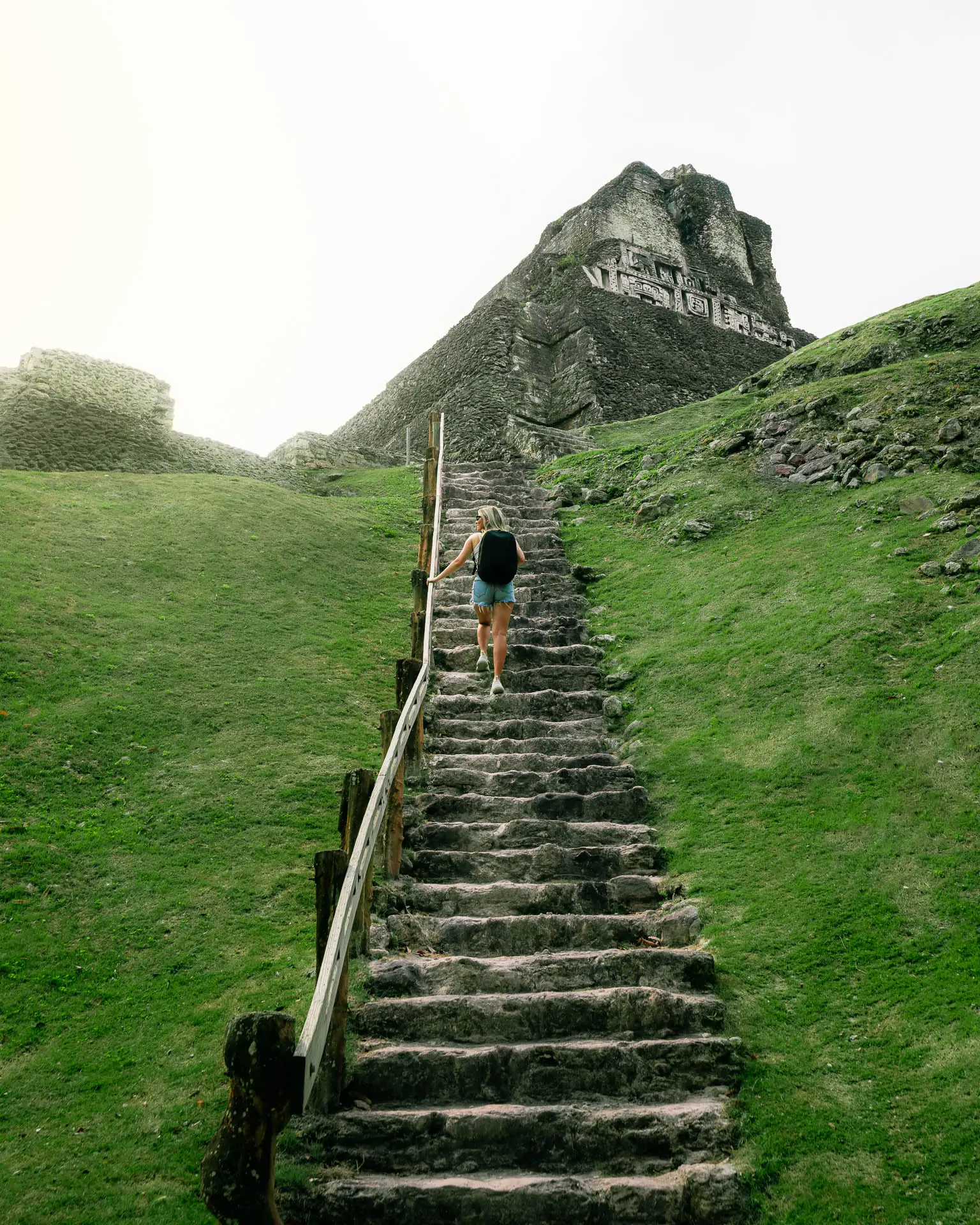 Person with backpack climbing stone steps toward the ancient Mayan pyramid Xunantunich surrounded by grassy hills.