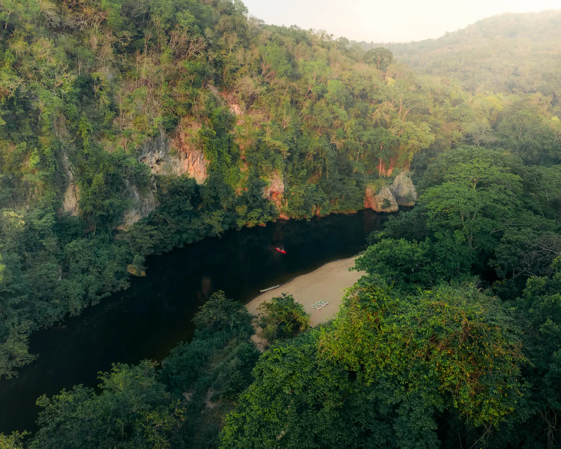 The Macal River winds through dense green jungle with a small sandy beach and a red canoe on the water at Sweet Songs Jungle Lodge in Belize.