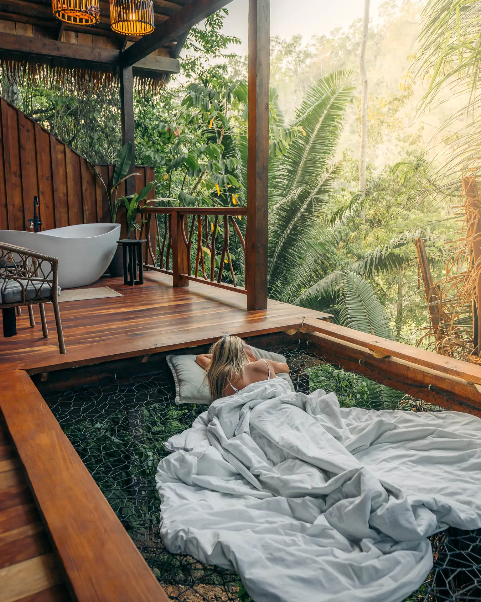 Woman lying on a netted hammock bed with white sheets on a wooden balcony overlooking lush jungle greenery at Sweet Songs Jungle Lodge in Belize.