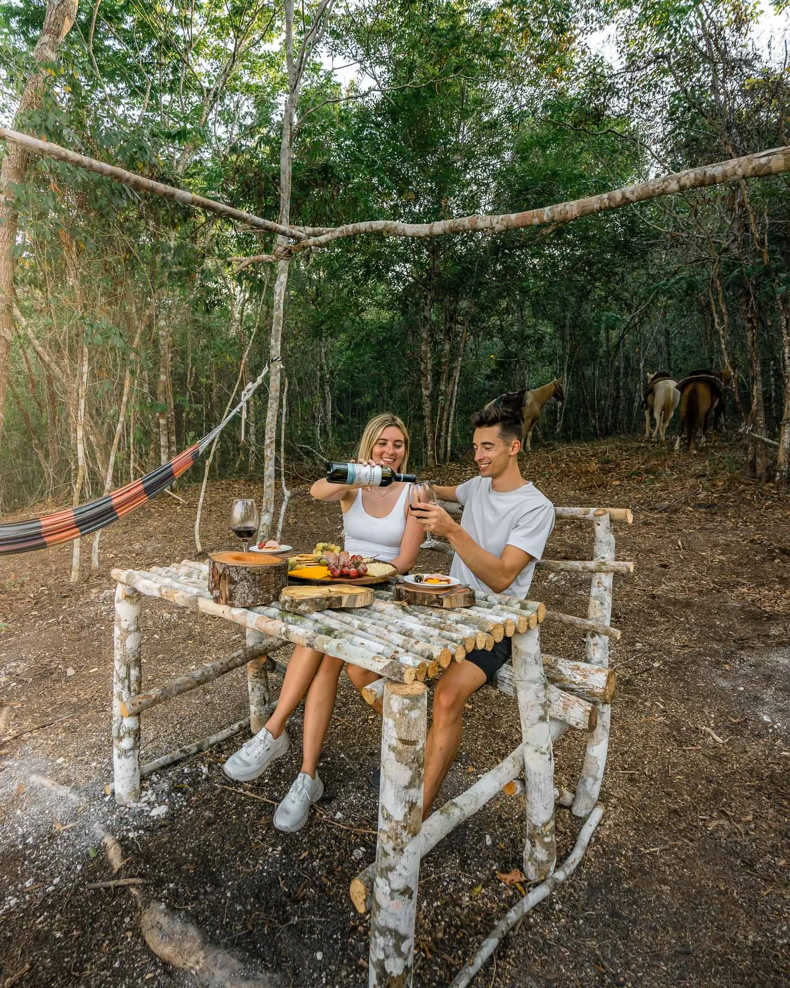 Couple sitting on rustic wooden bench at outdoor table in forest, pouring wine and enjoying snacks at Sweet Songs Jungle Lodge in Belize.