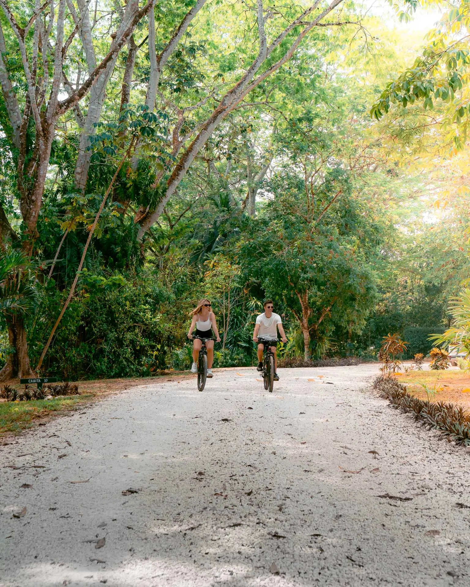 Two people cycling on a gravel path surrounded by lush green trees and foliage at Sweet Songs Jungle Lodge in Belize.