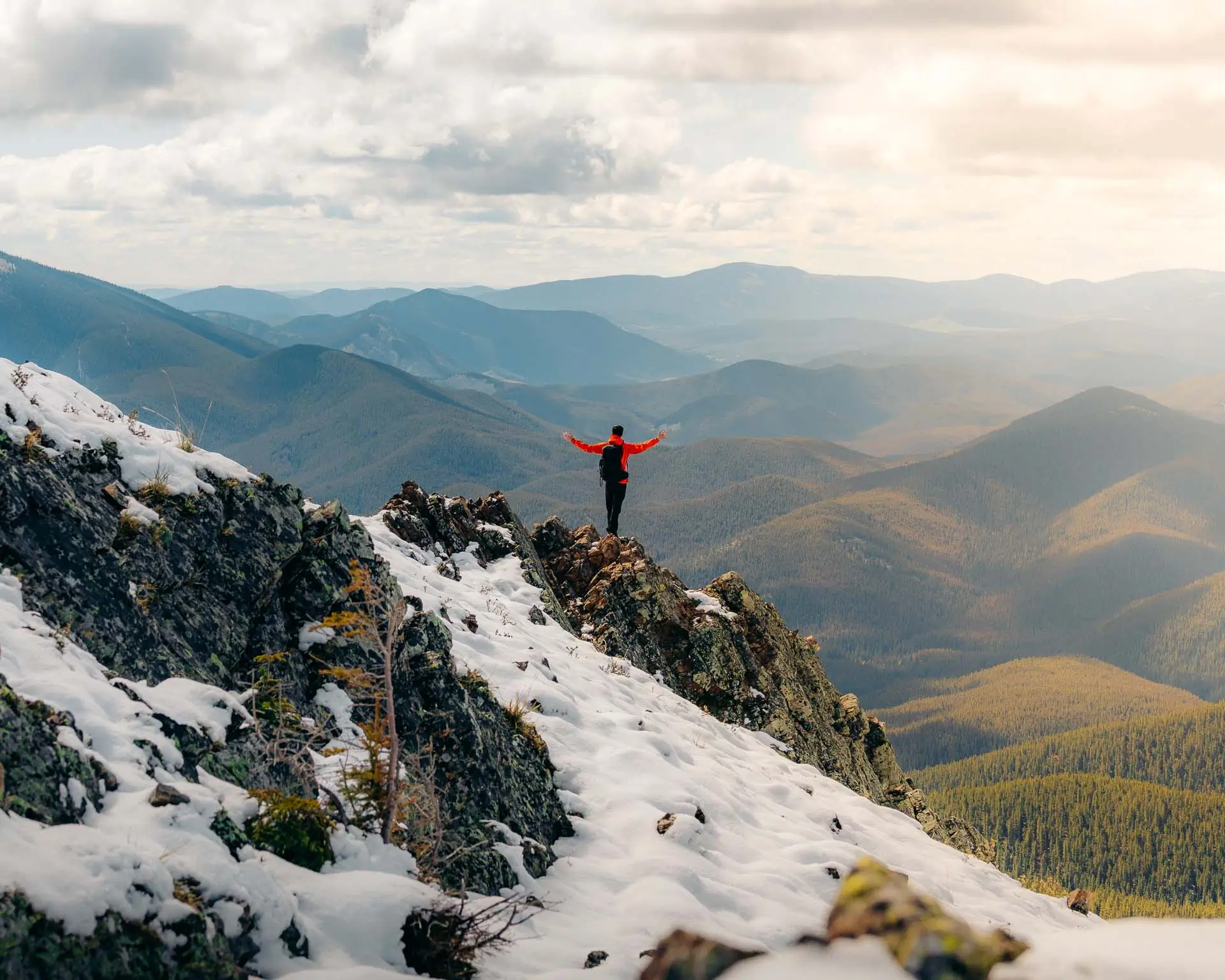 Hiker in red jacket celebrating with arms raised on a rocky ridge at the top of Jumpingpound Summit Trail in Kananaskis, Alberta.