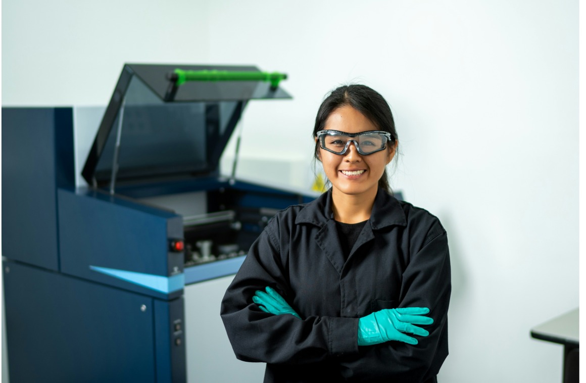 Woman in safety goggles and teal gloves standing in a lab in front of open equipment