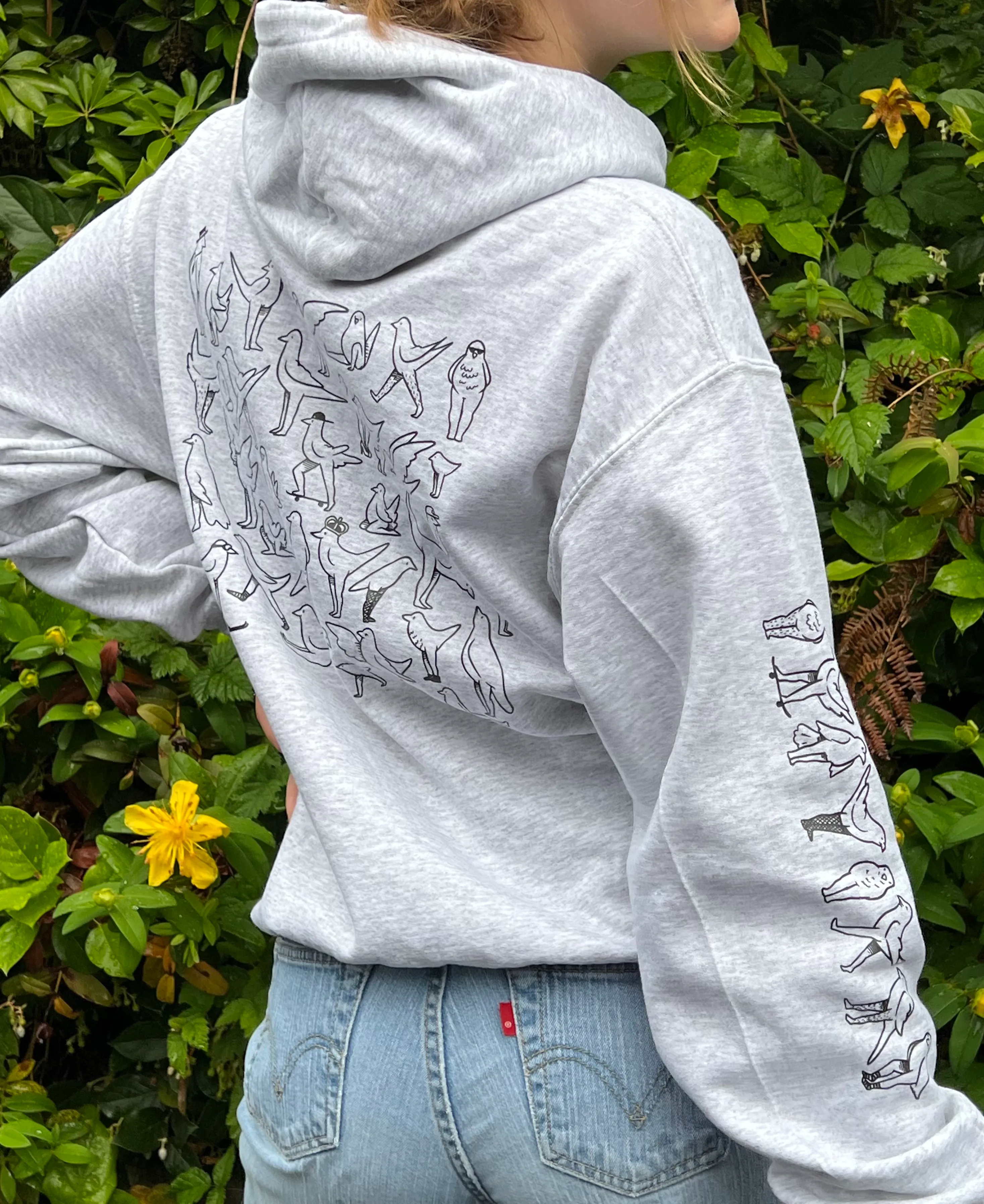Person wearing a light gray hoodie with black line drawings of various birds on the back and down the right sleeve, standing in front of green foliage with yellow flowers.