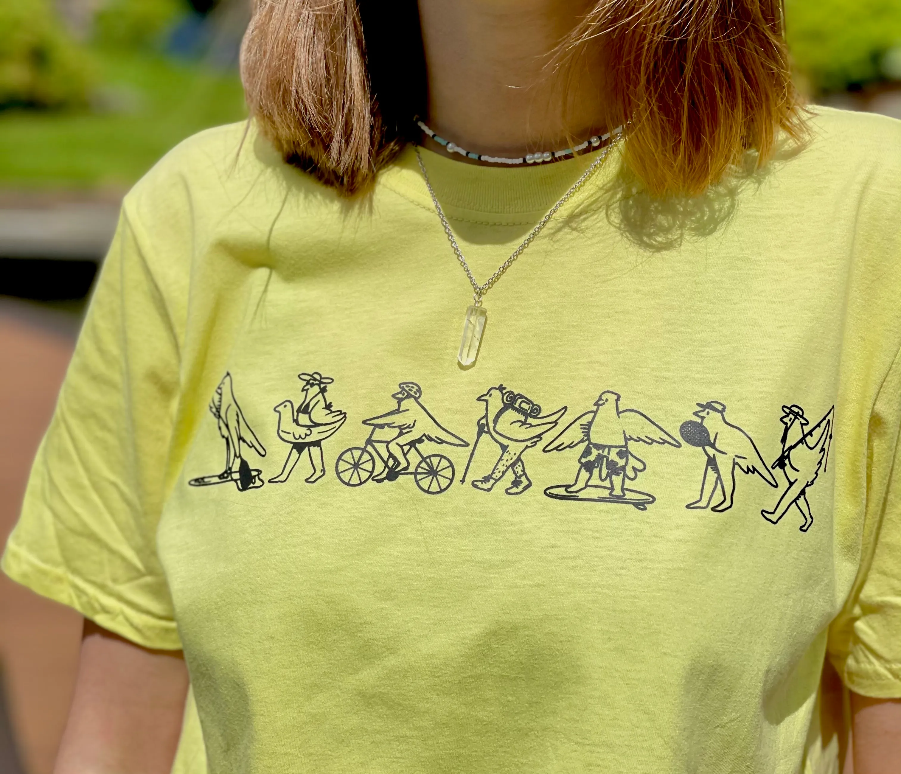 Yellow T-shirt with black line drawings of birds engaged in various activities like playing guitar, cycling, hiking, skateboarding, holding a ball, and fishing.