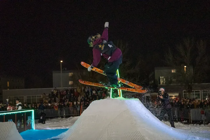 Skier performing a trick mid-air over a snow ramp at night during a ski event with a crowd watching.