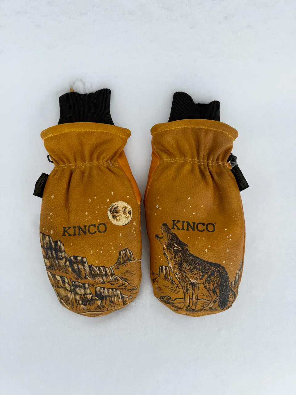 Pair of brown Kinco mittens on snow with a moon and canyon design on the left mitten and a howling wolf and canyon design on the right mitten.