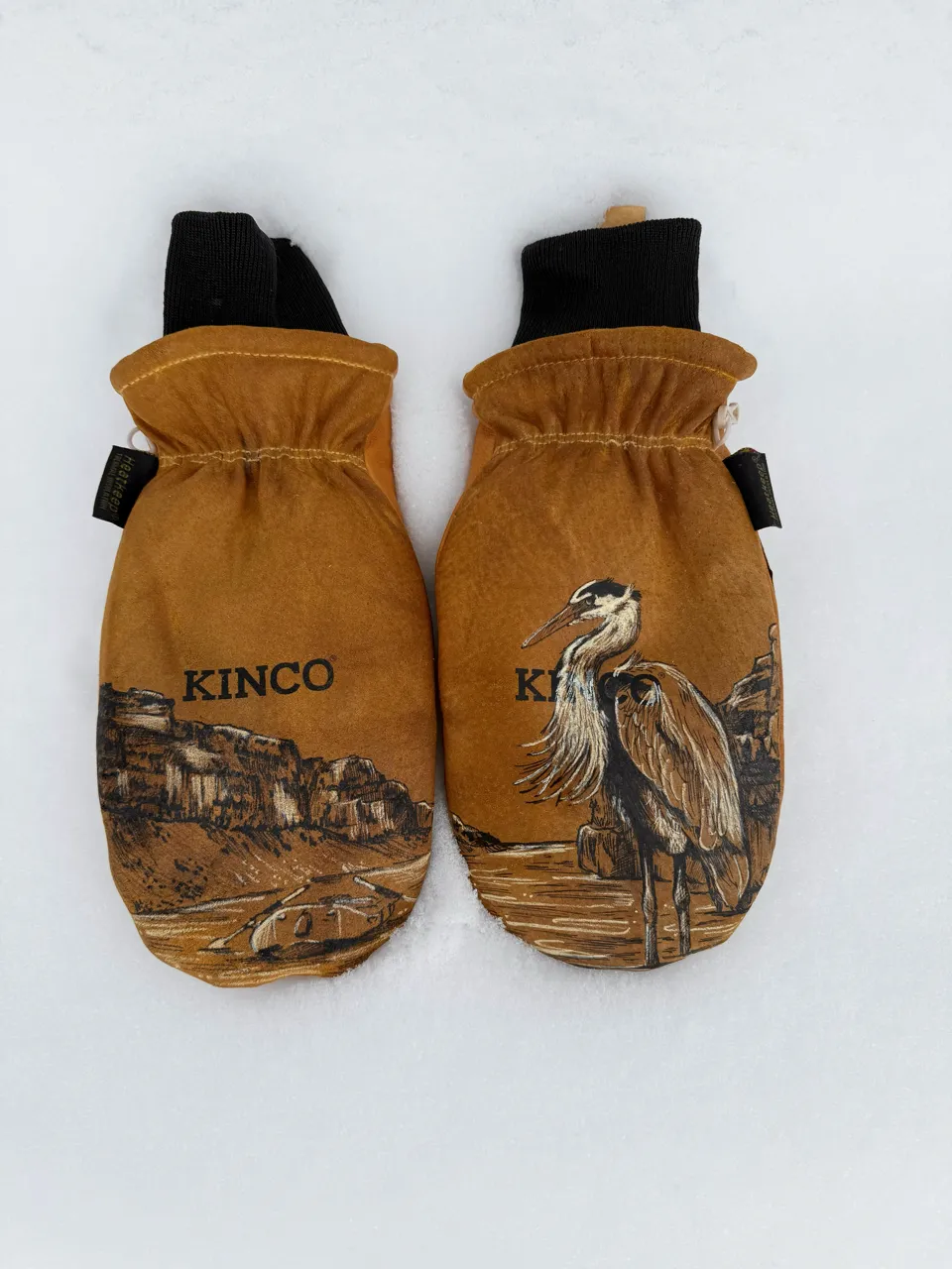 Pair of brown Kinco mittens with black cuffs, one mitten featuring a river and canyon scene, the other showing a heron bird illustration, lying on snow.