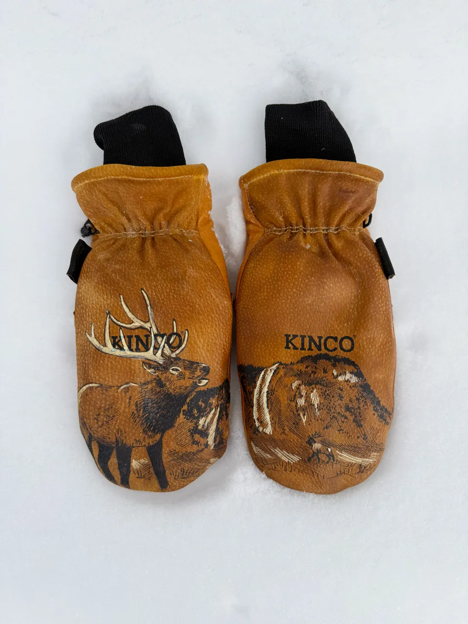 Pair of brown Kinco leather mittens with wildlife and mountain designs, lying on snow.