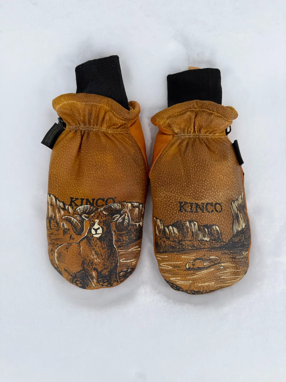 Pair of brown Kinco leather mittens with black cuffs, one showing a ram and the other a desert landscape, lying on snow.