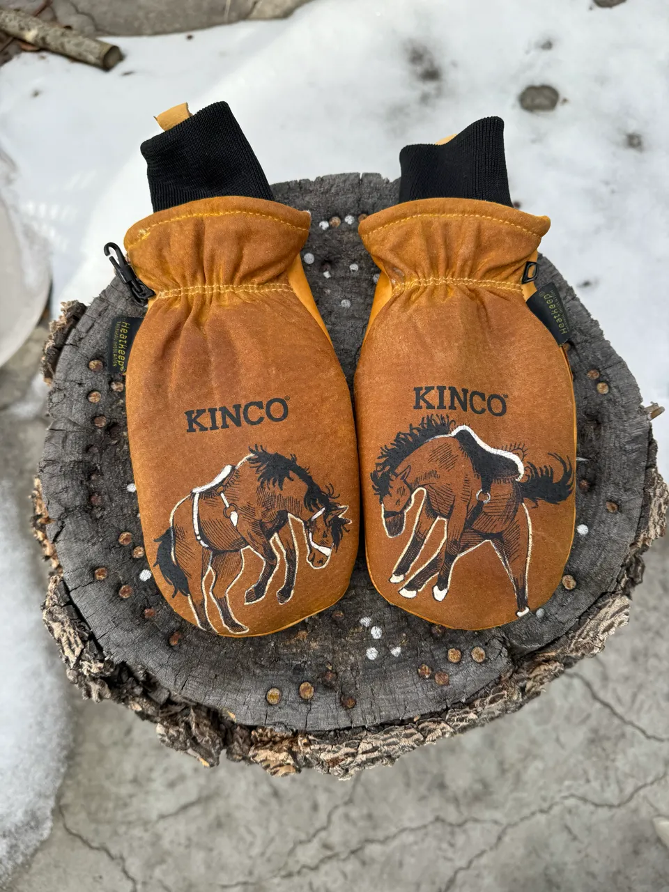 Pair of brown Kinco mittens with horse illustrations placed on a weathered tree stump against a snowy and concrete background.