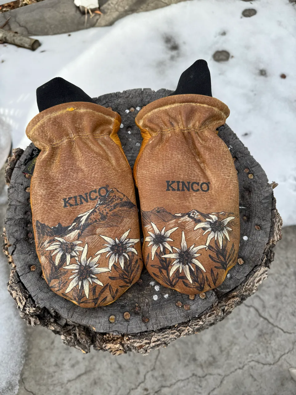 Pair of brown Kinco leather mittens with floral and mountain designs, resting on a tree stump with snow and concrete in the background.