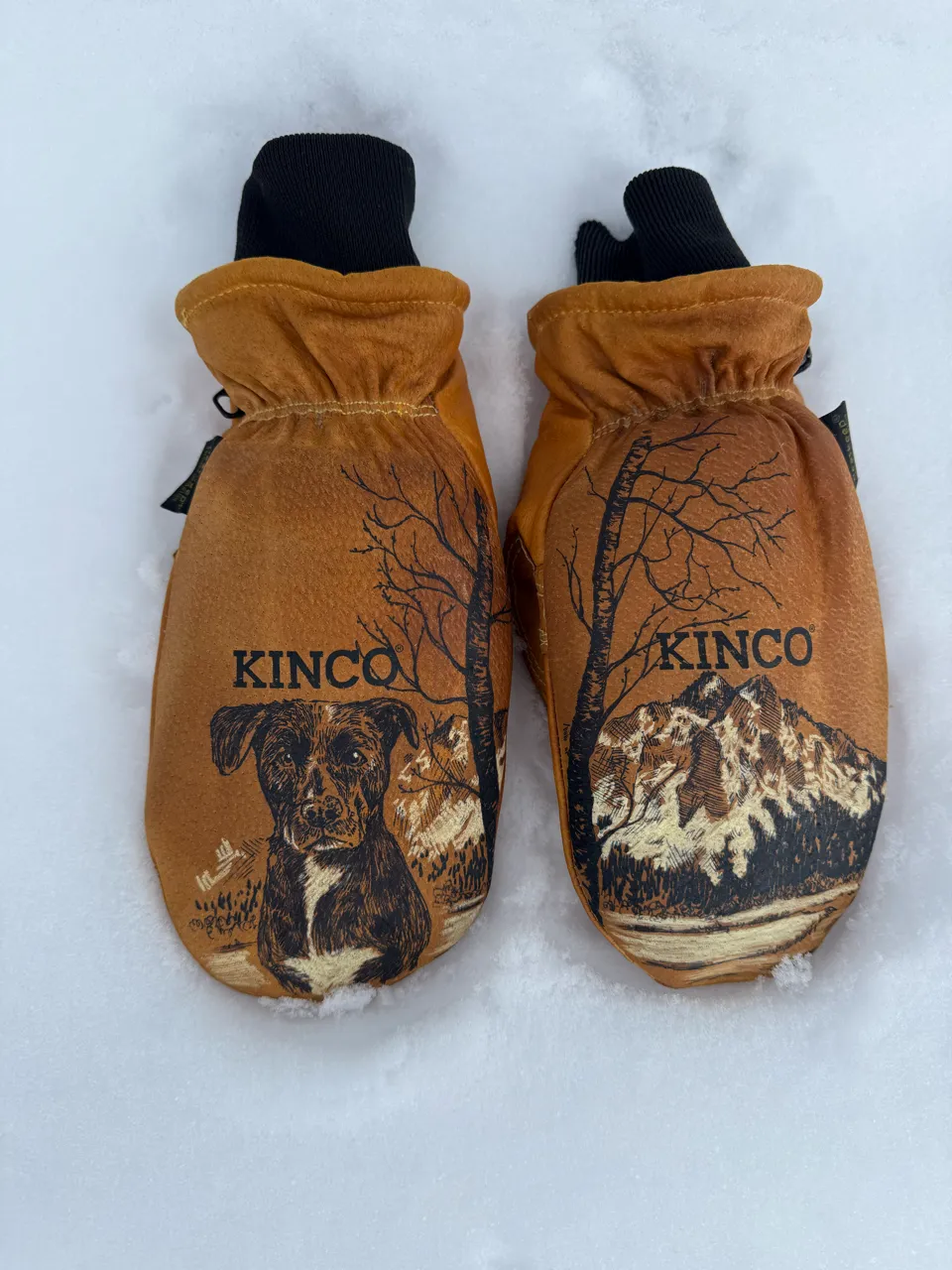 Pair of brown Kinco mittens with black cuffs, one featuring a black dog illustration and the other a mountain landscape, placed on snow.