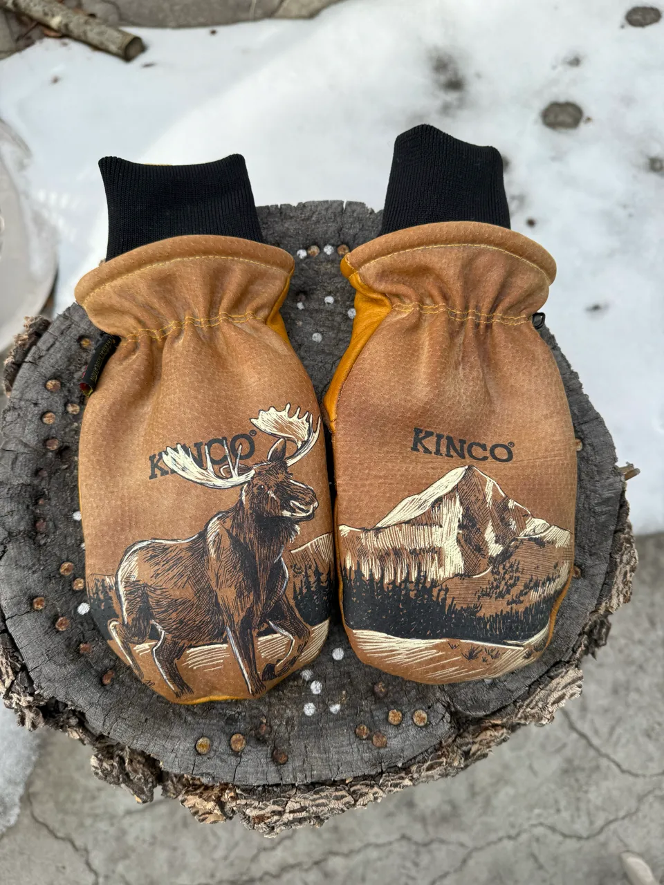 Pair of brown Kinco mittens with black cuffs, one featuring a moose illustration and the other a mountain landscape, placed on a tree stump with snow in the background.