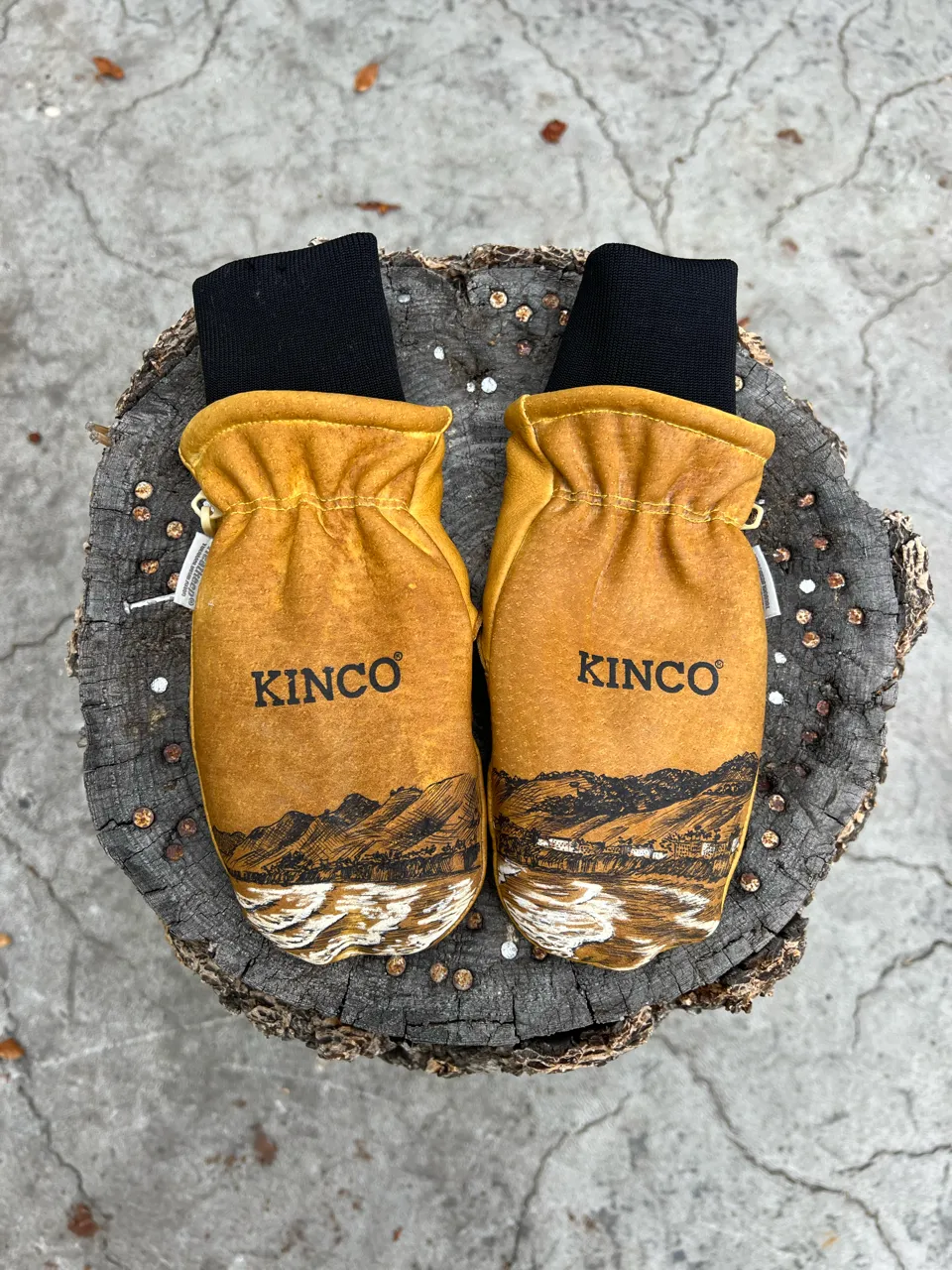 Pair of tan Kinco work gloves with black cuffs featuring a mountain and river landscape illustration, placed on a tree stump.