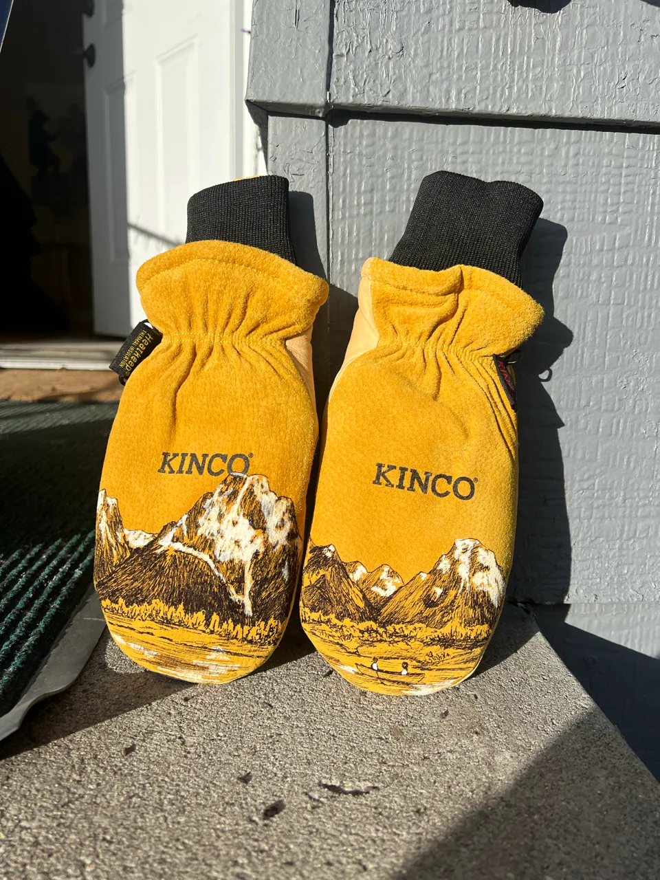 Pair of yellow Kinco work gloves with mountain landscape illustrations on the palms, leaning against a gray wooden wall.