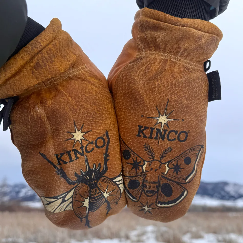Pair of brown leather mittens with black and white insect illustrations and the word 'KINCO' on each mitten, worn against a snowy mountain background.