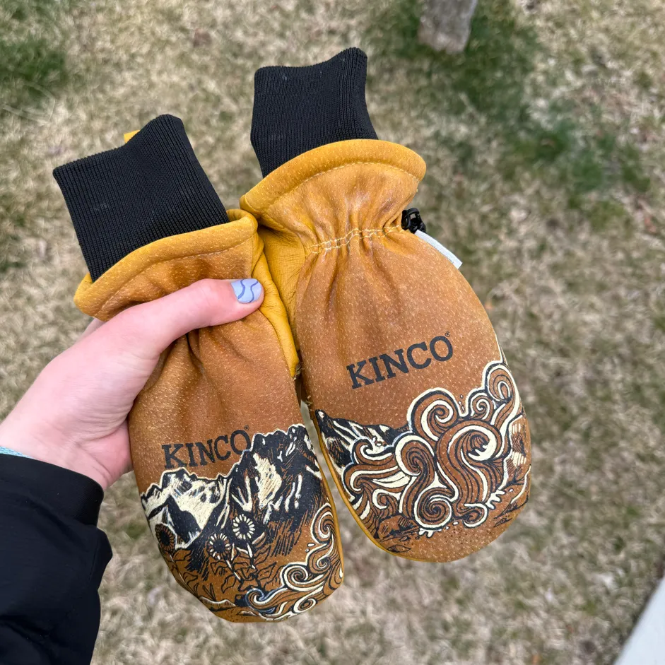 Hand holding a pair of brown Kinco mittens with mountain and floral designs on a grassy background.