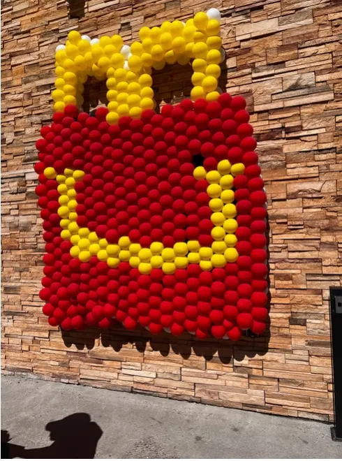 Large red and yellow balloon sculpture mounted on a stone wall, resembling a tote bag with yellow handles and a yellow smiley face design.