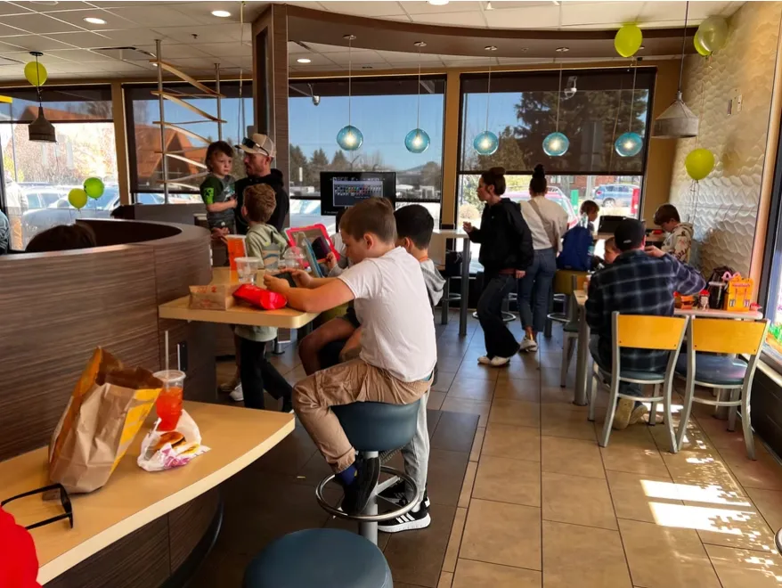 Busy fast food restaurant interior with children and adults sitting and standing, illuminated by hanging globe lights and natural daylight.