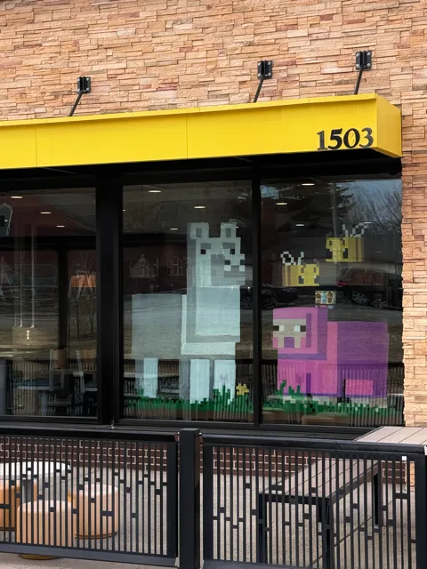 Shop window with pixel art of a white llama, a pink sheep, and two yellow bees on a stone brick building with a yellow awning numbered 1503.