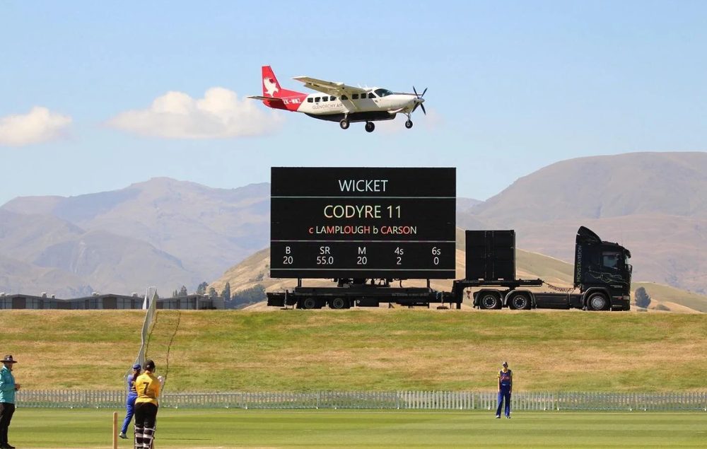 Mobile LED scoreboard truck at a cricket ground with mountains behind