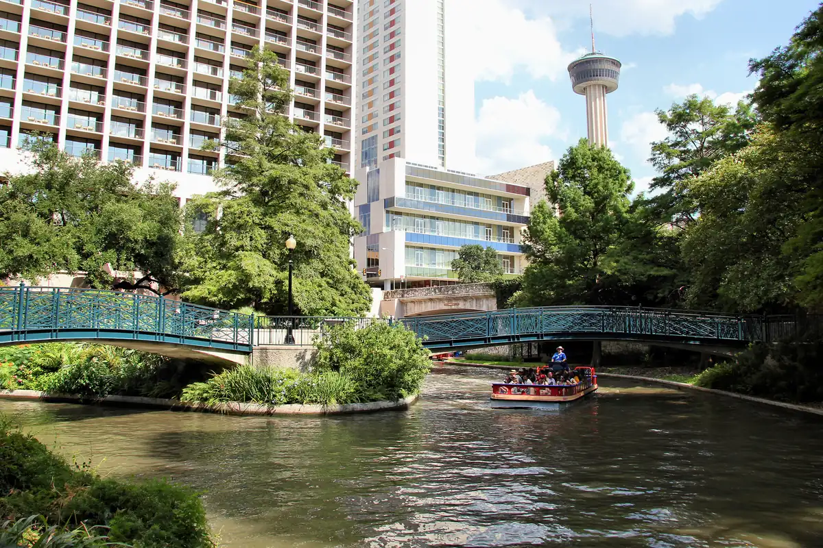 The San Antonio River Walk lined with trees, with the Tower of the Americas rising in the background