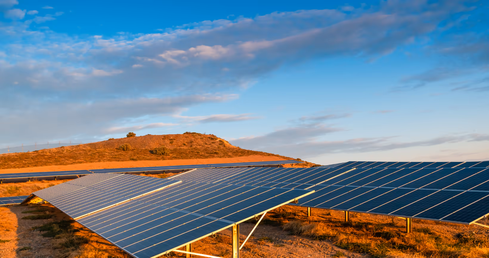 Large-scale solar farm installation in desert landscape at sunset with red rock hill in background