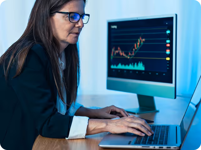 Female professional with glasses analyzing financial data charts on multiple computer monitors while working on laptop